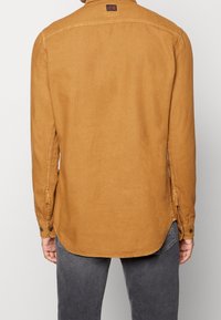 Brązowa koszula z długim rękawem z zaokrąglonym dołem, mankietami na guziki i bocznymi rozcięciami. Wykonana z tkaniny o fakturze, z małą metką przy dekolcie.