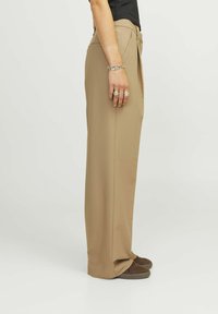 Pantalons larges marron clair avec une texture lisse, présentant une coupe décontractée et un devant plissé, associés à des chaussures marron foncé.