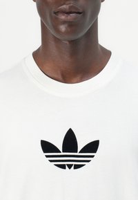 Witte katoenen t-shirt met een opvallend zwart Adidas trefoil logo en drie horizontale strikken onder het logo op de borst.