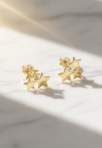 Des boucles d'oreilles en forme d'étoile dorée présentent trois étoiles de tailles variées avec une finition métallique lisse, projetant de douces ombres sur une surface claire.