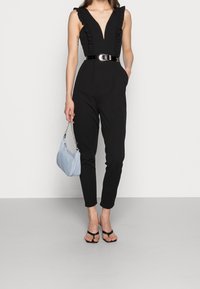 Mono negro con acentos de hombro con volantes, escote en forma de V profundo y cintura ajustada, complementado por un cinturón negro brillante y un bolso de mano azul claro.