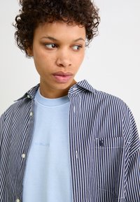 Chemise à rayures avec une poche, superposée à un sweat-shirt col rond bleu clair arborant un logo subtil. Tissu texturé et style décontracté.