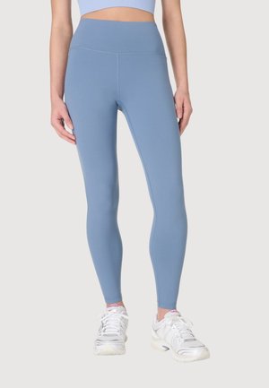 Vrouw draagt lichtblauwe legging met hoge taille en witte hardloopschoenen tegen een effen achtergrond.