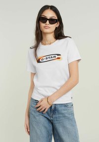 Bílý bavlněný tričko s grafickým logem "G-STAR" v černé a oranžové barvě. V kombinaci se světle modrými džíny model nosí černé sluneční brýle.