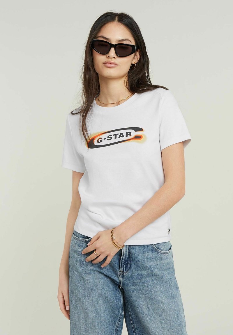 Bílý bavlněný tričko s grafickým logem "G-STAR" v černé a oranžové barvě. V kombinaci se světle modrými džíny model nosí černé sluneční brýle.