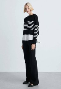 Gestreifter Strickpullover in Schwarz-Weiß, über einem weißen Hemd getragen, kombiniert mit schwarzen, weit geschnittenen Hosen und schwarzen Ballerinas. Minimalistisches Design.