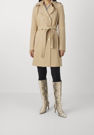 Femme portant un trench-coat beige ceinturé sur un pantalon noir et des bottes à talons hautes jusqu'au genou avec un motif peau de serpent, debout sur un fond neutre.