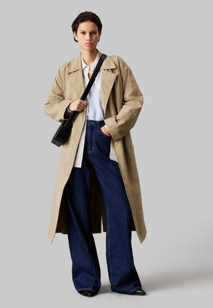 Vrouw draagt een beige trenchcoat, wit overhemd, brede blauwe spijkerbroek, zwarte schoenen en draagt een zwarte crossbodytas, staand tegen een grijze achtergrond.