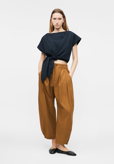 Γυναίκα όρθια με τα χέρια στις τσέπες, φορά μαύρο κοντό τοπ δεμένο, φαρδιά καφέ παντελόνια και μαύρα επίπεδα παπούτσια, μπροστά σε απλό ανοιχτό φόντο.