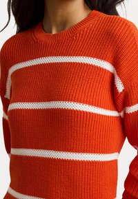 Stickad tröja i klar orange med vita horisontella ränder. Har rund halsringning och texturerat tyg. Nära bild av designen.