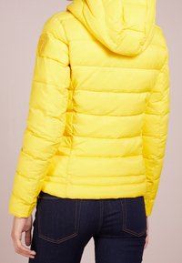 Giacca a vento gialla con cappuccio, caratterizzata da un design trapuntato e una texture imbottita. Include una toppa con logo sulla parte superiore del braccio e una silhouette aderente.
