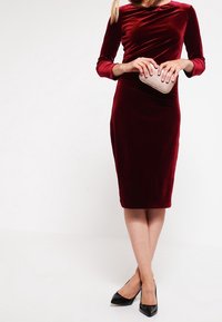 Robe en velours bordeaux à manches longues, design ajusté, longueur sous le genou, accompagnée d'une pochette texturée et de talons noirs.