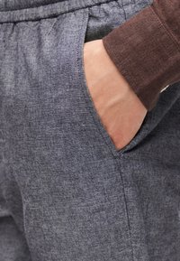 Χέρι στην πλαϊνή τσέπη μπλε υφασμάτινου παντελονιού, με καφέ πουκάμισο μακριάς μανίκας.