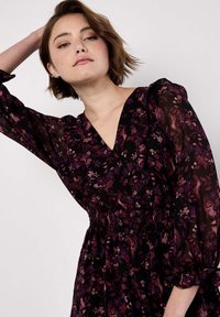 Schwarzes Kleid mit einem floralen Muster in Lila- und Bordeauxtönen; mit V-Ausschnitt und Puffärmeln; tailliert mit Smok. 