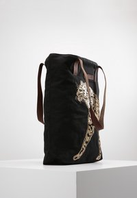 Sac fourre-tout en toile noire avec des anses en cuir marron et un motif léopard en sequins dorés sur le côté. Forme rectangulaire, tissu texturé.