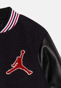 Černá univerzitní bunda s vlněným tělem a koženými rukávy. Obsahuje červené vyšívané logo Jumpman a pruhovaný ribbed límec.