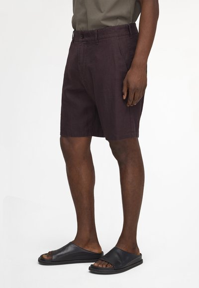 Homme portant un short violet foncé jusqu'aux genoux et des sandales noires à bout ouvert, debout devant un fond blanc uni.