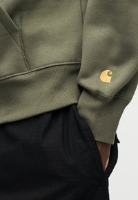 Φόρμα σε ελιά πράσινο με ριμπ μανσέτες και τελείωμα, διακοσ μημένη με ένα μικρό χρυσό λογότυπο στο αριστερό μανίκι. Κάτω φαίνονται σκουρόχρωμοι υφασμάτινοι παντελόνες με υφή.