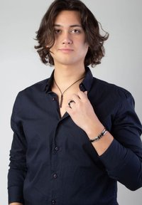 Zwarte langslavige shirt met knopen, zilveren hangerketting, een ring en een zwarte armband, gedragen door een persoon met golvend haar.