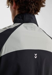 Svart och ljusgrå jacka med hög krage, geometrisk färgblockering och subtilt chevrondesign på baksidan. Mjuk, slät tekstur.