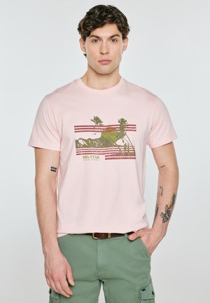 Mężczyzna w różowym t-shircie z zielonym wzorem roślinnym i napisem "BIG STAR", w zielonych spodniach i brązowym pasku, stoi z ręką w kieszeni.