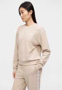 Μπεζ φούτερ με ριμπ λαιμόκοψη και μανσέτες, με διακριτικό λογότυπο EA7 στο στήθος. Μαλακό υλικό με άνετη εφαρμογή, φορεμένο με ταιριαστά παντελόνια.