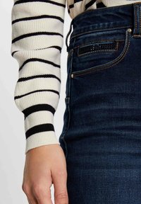 Jeans de tiro alto de mezclilla oscura con una textura suave, costuras delineadas y bolsillos visibles. Combinados con un suéter a rayas blanca y negra.