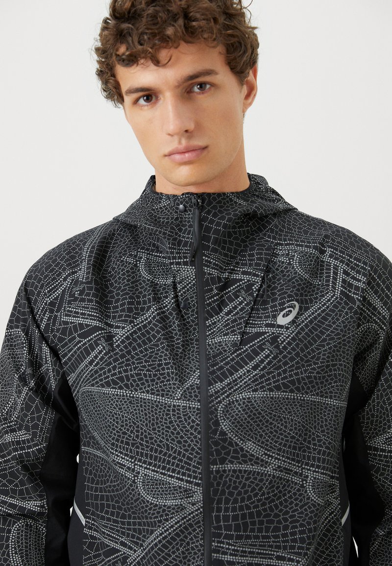 Chaqueta negra con un patrón geométrico texturizado en plata. Incluye un cierre de cremallera y capucha. El material parece ligero y transpirable.