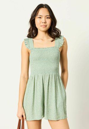 Mladá žena s tmavými vlasmi po plecia, oblečená v svetlozelený overal bez rukávov s volánovými ramienkami, ktorá drží hnedú kabelku.