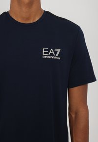 Námorně modré bavlněné tričko s kulatým výstřihem, na levém prsu obsahuje logo EA7 v bílé barvě a texturované černé akcenty na ramenech.