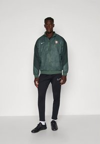 Grønn bomberjakke med glidelås og strukturert mønster, hvit Nike-logo, svarte bukser med Nike-logo og svarte treningssko.