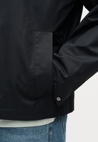 Άτομο που φοράει μαύρο μπουφάν με το χέρι στην τσέπη στο πλάι, το κουμπί στη μανσέτα εμφανές, μπλε τζιν και ανοιχτόχρωμο πουκάμισο από κάτω.