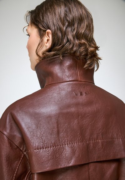 Personne aux cheveux courts ondulés bruns porte une veste en cuir marron à col haut avec "VSP" embossé dans le dos.
