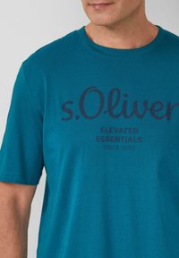 Türkisfarbenes T-Shirt aus Baumwolle mit kurzen Ärmeln. Auf der Vorderseite befindet sich der dunkelblaue Drucktext "s.Oliver ELEVATED ESSENTIALS SINCE 1969".