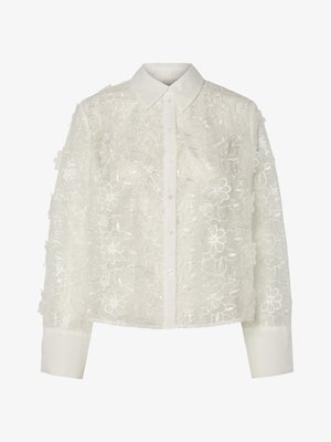 Valge pluus lillelise pitskattega, millel on krae, nööbid ees ja pikad varrukad. Struktuurne toime tõstetud tikitud lilledega.