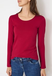 T-shirt a maniche lunghe e collo rotondo in cotone rosso. Presenta una silhouette aderente e una texture liscia. Abbinato a jeans in denim blu.