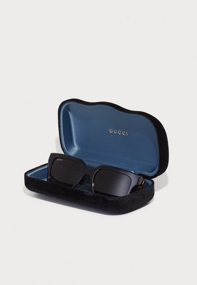 Sorte solbriller i et fløyelsfôret etui med blå innside. Etuiet har et bølgete design; solbrillene har mørke linser og gullfester.