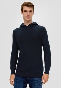 Junger Mann in einem enganliegenden, dunkelblauen Strickhoodie und blauen Jeans, der vor einem schlichten, hellgrauen Hintergrund steht.