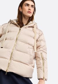 Beige pufferjack met een capuchon, voorzien van diagonale quilting, gouden hardware en verstelbare banden. Ritszakken op de mouwen.