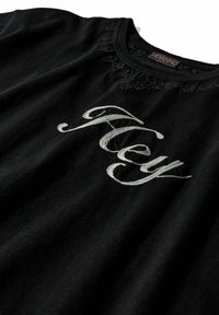 Camiseta de algodón negra con cuello redondo, delicado borde de encaje y texto bordado en plata que dice "Hola". Textura suave.