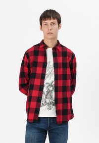 Jauns vīrietis, kurš valkā sarkanu un melnu četru stūrīšu krekliņu virs balta grafiskā T-krekla ar palmu attēliem un zilu džinsu, stāv pret vienkāršu fona.