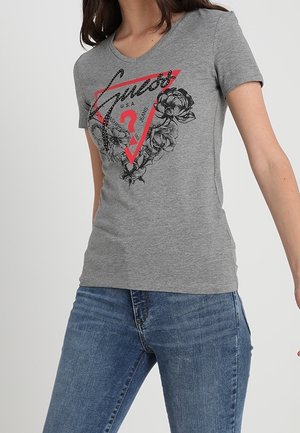 Woman wearing a fitted grey t-shirt with a red triangle, black floral design, and the word "Guess" paired with blue jeans.