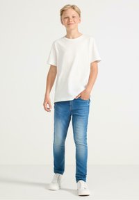 Witte t-shirt met korte mouwen, blauwe slim-fit jeans en witte sneakers. Lichte denim met subtiele vervaging, casual ontwerp. Staande pose.