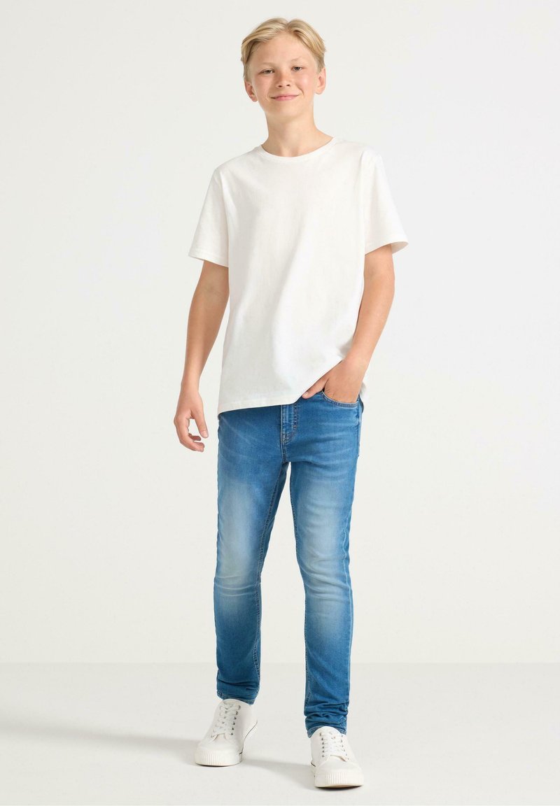 Witte t-shirt met korte mouwen, blauwe slim-fit jeans en witte sneakers. Lichte denim met subtiele vervaging, casual ontwerp. Staande pose.