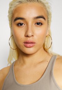 Modelo com cabelo liso e loiro claro, usando grandes brincos de argola dourados e uma camiseta regata canelada de um tom bege claro. Maquiagem natural, expressão neutra.