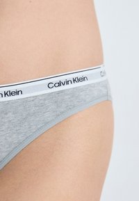 Szürke pamut tanga fehér elasztikus derékvonallal, amelyen fekete "Calvin Klein" márkajelzés található. Simává formázott textúra és minimalista dizájn.