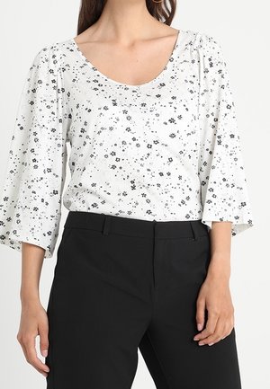 Femme portant un chemisier blanc à petit motif floral noir et manches cloche 3/4, associé à un pantalon noir taille haute.