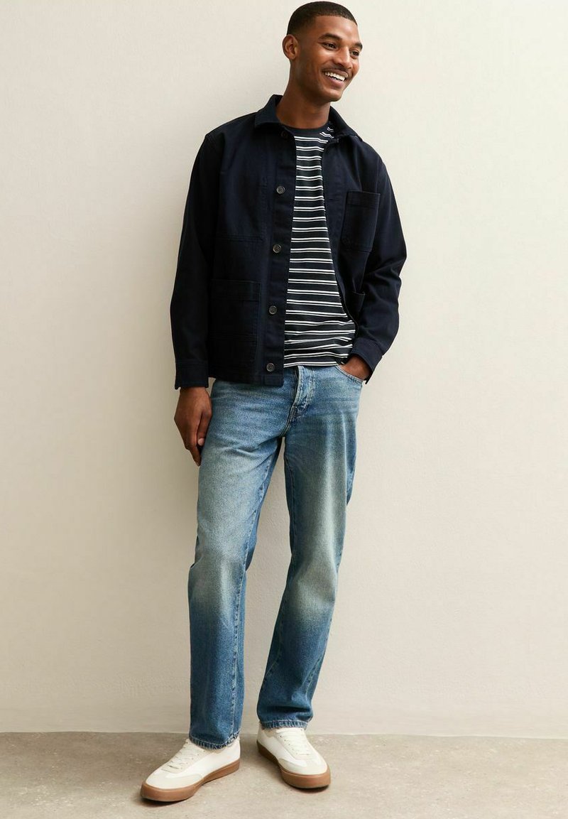 Marineblauwe jas over gestreepte T-shirt, gecombineerd met lichtblauwe jeans en witte sneakers met tan zolen. Casual, ontspannen pasvorm met knoopdetails.