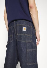 Dark blue denim work pants with multiple pockets, contrast yellow stitching, and a square Carhartt logo patch on the back pocket.