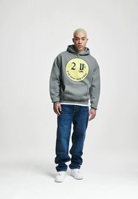 Grå hettegenser med en sirkulær gul grafikk som viser «2Y» og «f#k your bad vibes», kombinert med blå jeans og hvite joggesko.